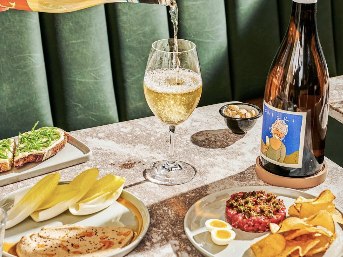 A spread of small plates such as charred eggplant yogurt, steak tartare, and cheese and charcuterie while a glass of sparking wine is poured.