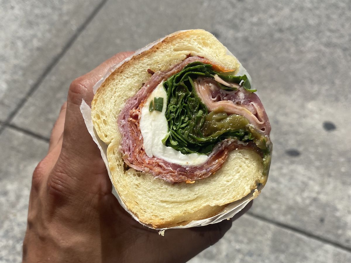 A hand clutches an Italian sandwich wrapped around itself with meat, cheese, and lettuce.