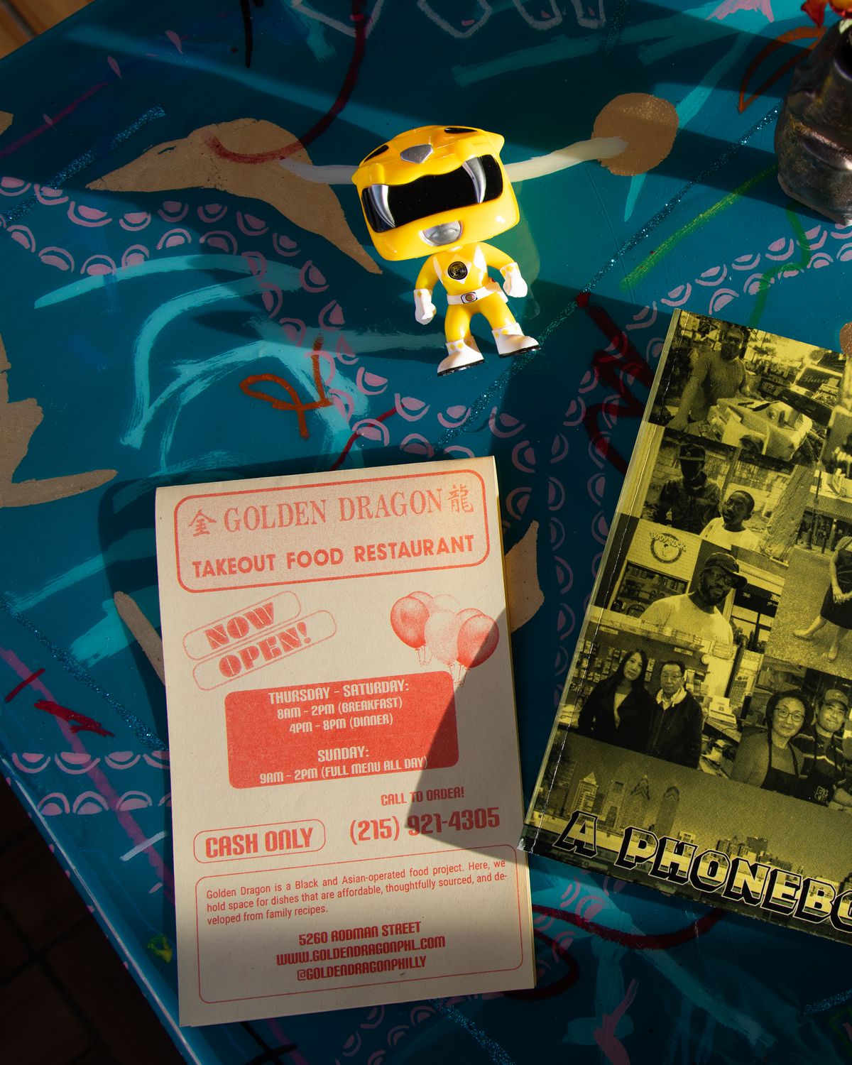 An overhead shot of a menu that reads Golden Dragon Takeout Food Restaurant, a yellow power ranger toy, and a black and white zine with yellow overlay.