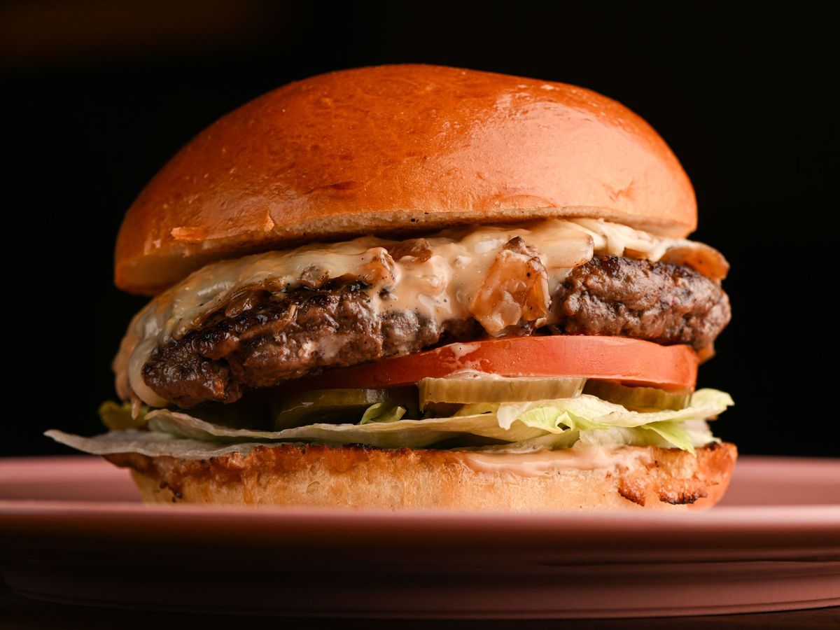 A close up side shot of a cooked burger with melted cheese and toppings at the bottom, against a black background, at restaurant Charcoal.