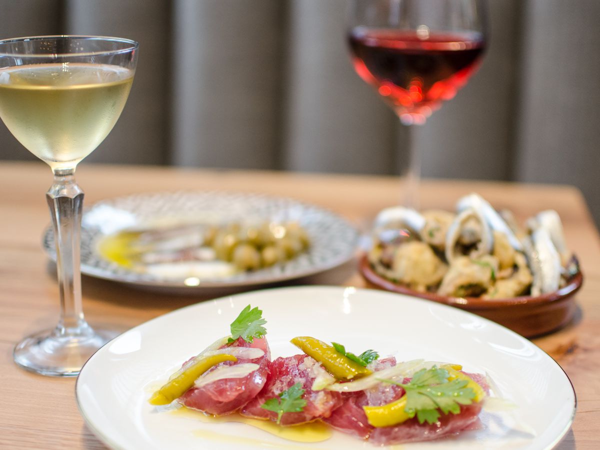 Three small tapas dishes sit on a light wooden table with a glass of white wine and red wine visible. The dish in the foreground is a tuna crudo, while the other two dishes are blurred in the background.