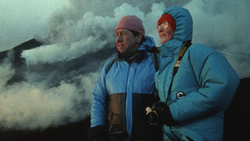 Two people in blue thermal jackets stand in front of a cloudy landscape.