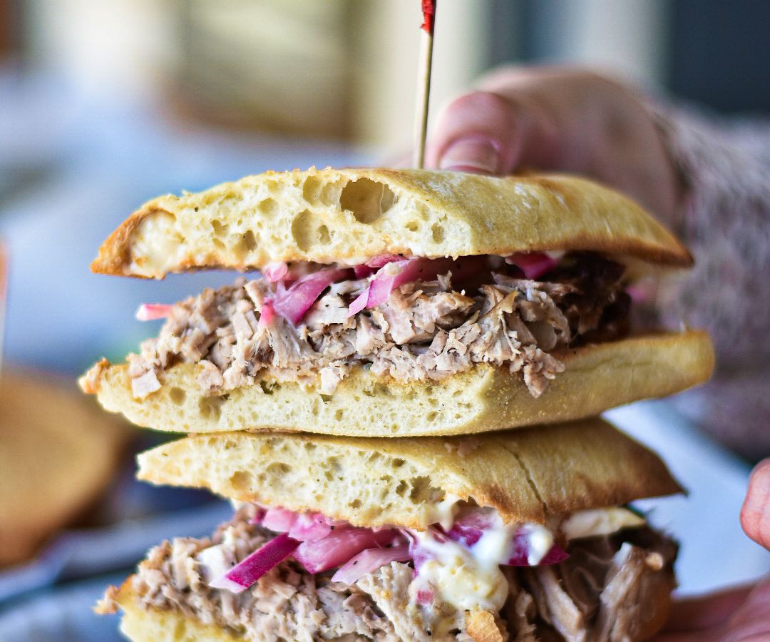Hands hold up two halves of a meaty sandwich dotted with purple cabbage 