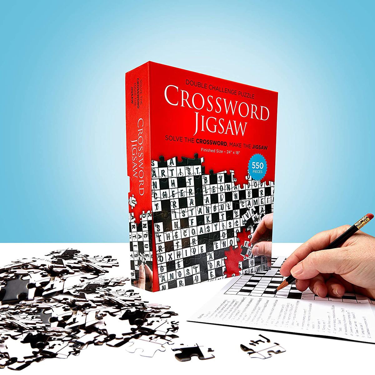 A hand works a crossword puzzle with a puzzle box and pieces in the background