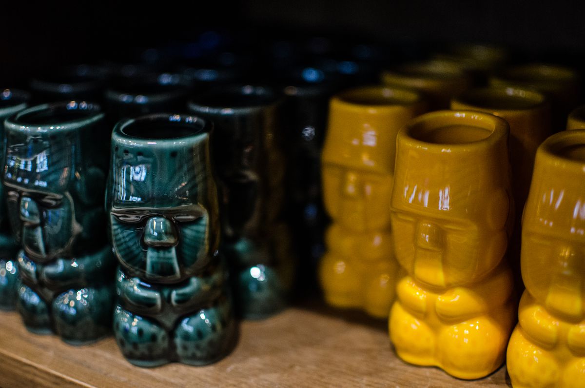 Small traditional tiki mugs, some green and some yellow, sit on a wooden shelf.