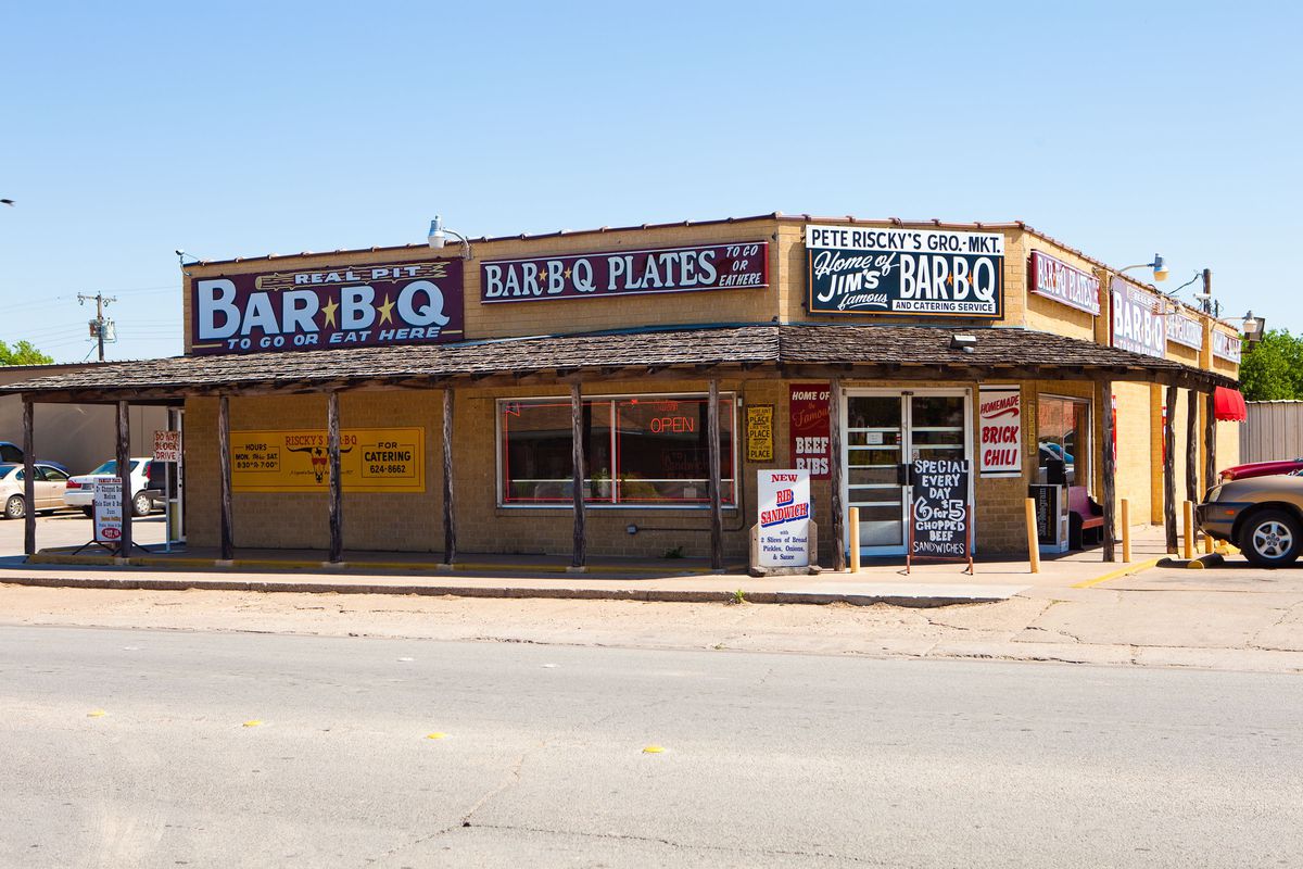 The orange/brown exterior of a barbecue restaurant, with multiple black and white signs advertising it’s food.