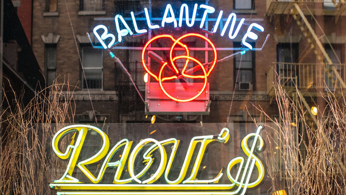 A neon sign with the words “Ballantine Raoul’s” gleams on a glass window.