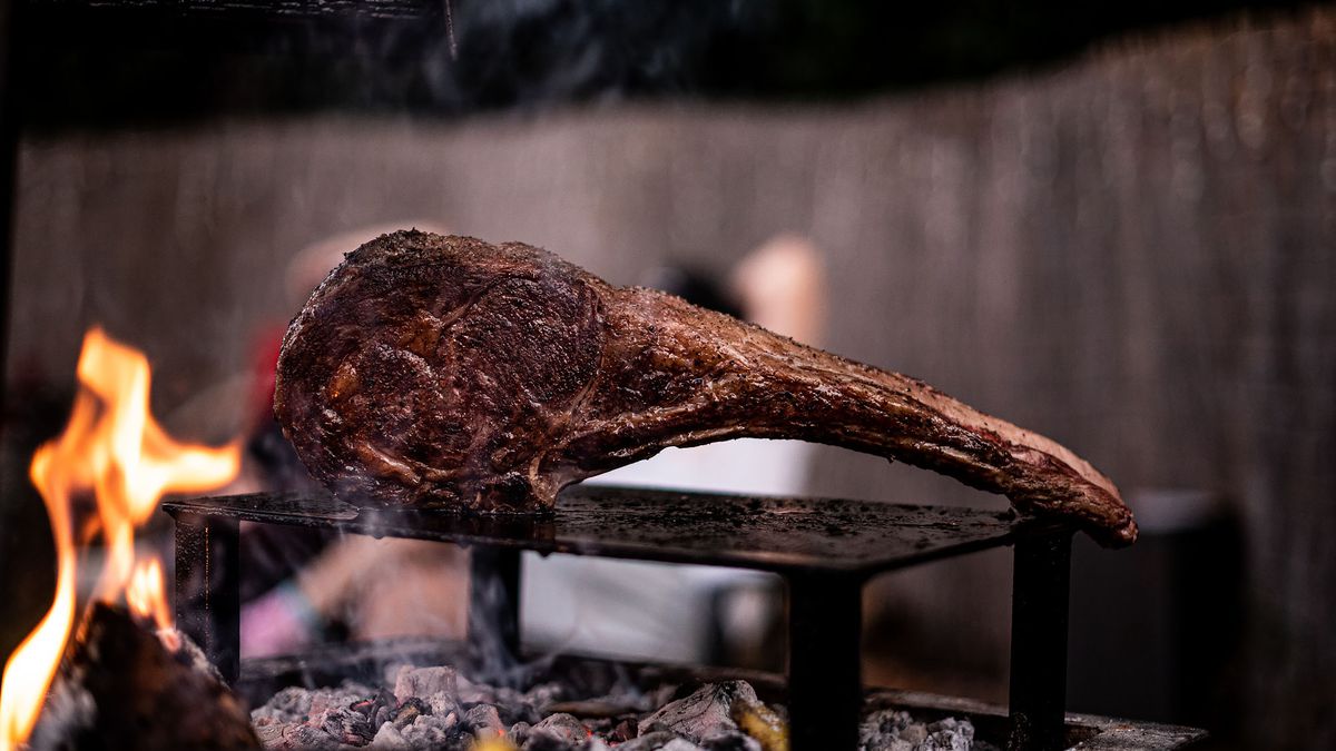 An evening photo of a steak shown on its side above a large flame.