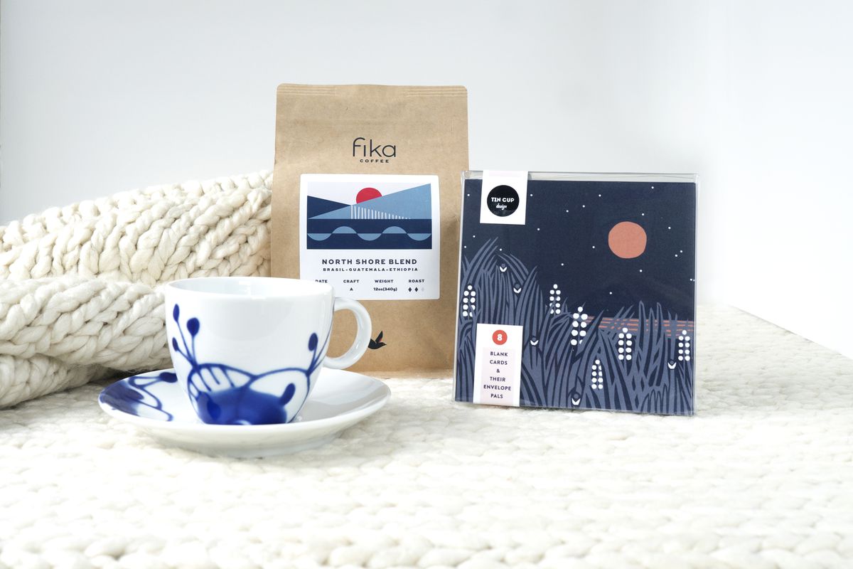 A white and blue tea cup in front of a bag of Fika coffee in a brown bag with Scandinavian designed label of blue and gray blue geometric shapes. A package of notecards are deep navy with white and gray wild rice stalks in front of a red harvest moon