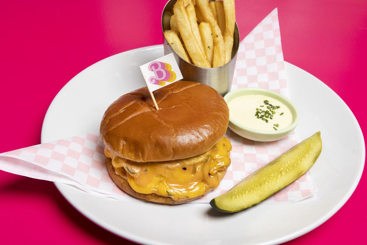 A round white plate with a cheeseburger, pickle spear, and metal cup of fries.