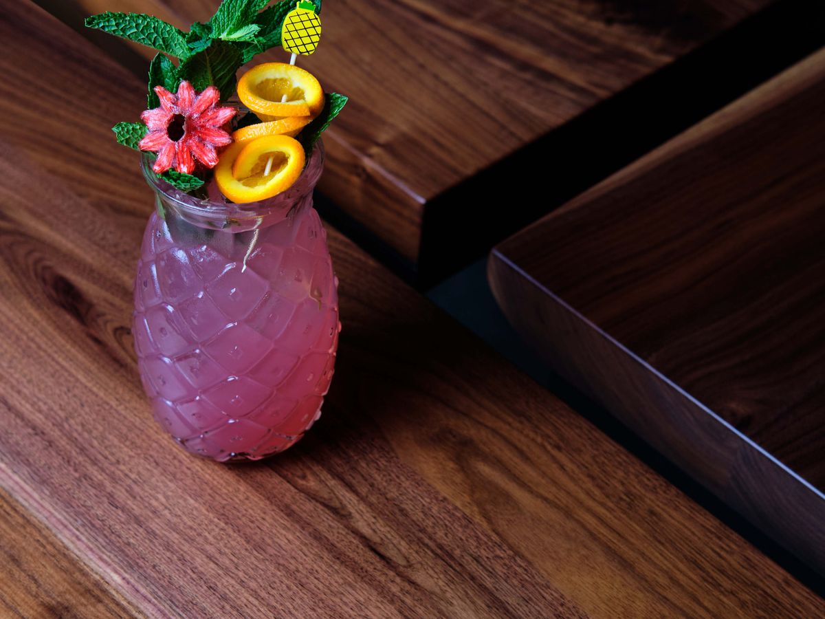 A bright pink and purple cocktail in a pineapple-shaped class, topped with mint, a pink flower, and circular orange peels hanging on a pineapple toothpick for a garnish.