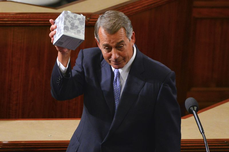 Boehner, clean shaven with his grey hair neatly cut, stands in front of a microphone on the House floor in a dark suit and purple tie. He holds a grey box of tissues aloft. 