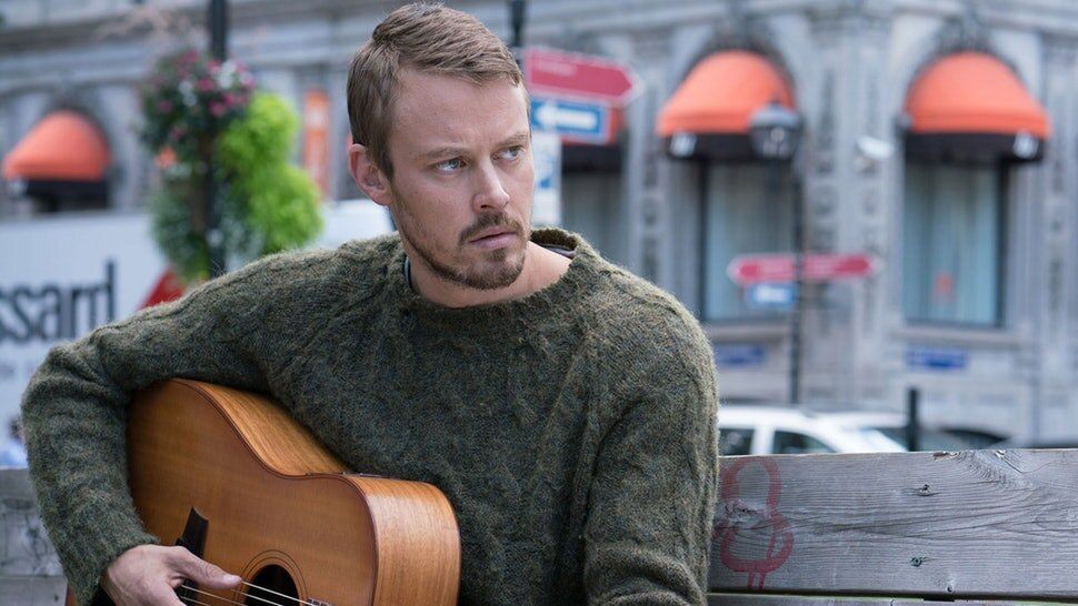Michael Dorman, wearing a sweater, sits on a park bench while playing a guitar in Patriot.