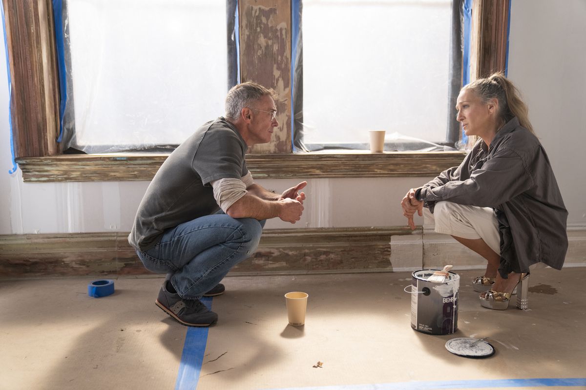 The characters Steve and Carrie in “And Just Like That.” They’re speaking, crouched on a floor, in a room that  looks like it’s being remodeled, with plastic on the windows and an open paint can on the floor. 