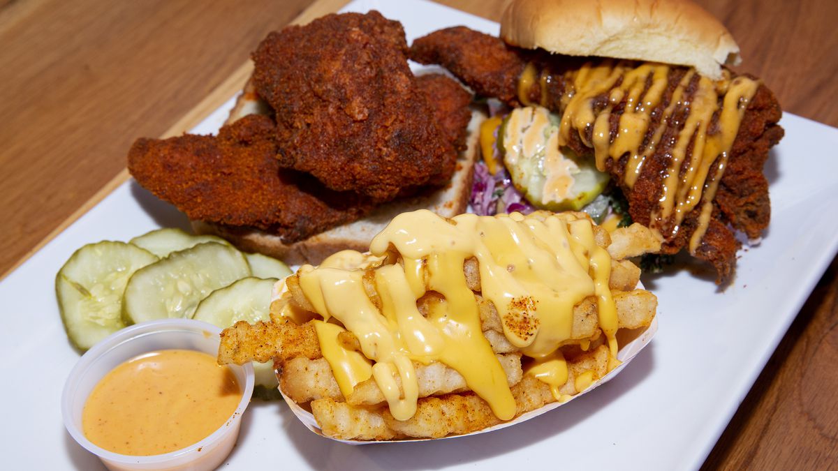 A white plate of two hot chicken sandwiches and a serving of crinkle-cut fries drizzled with cheese sauce.