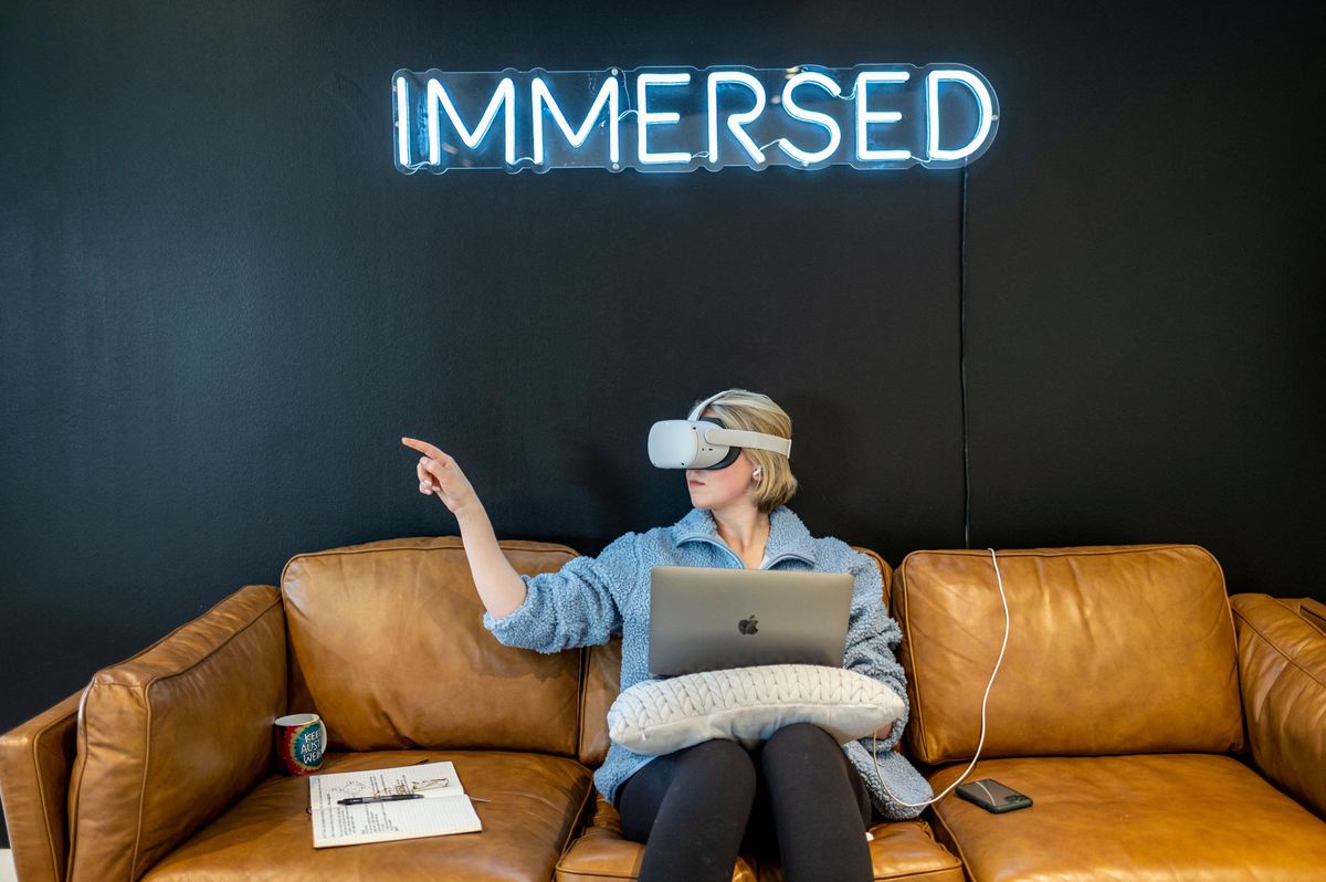 A person wearing a virtual reality headset sits with a laptop on a leather couch and points to one side. A neon sign on the wall behind spells out “immersed.”