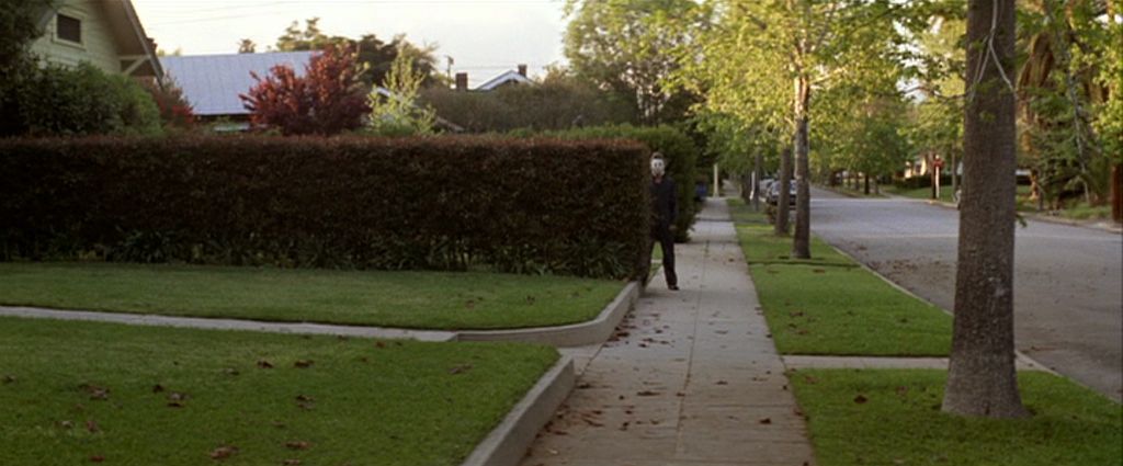 Michael Myers lurks near a hedge on a sidewalk in the original Halloween.