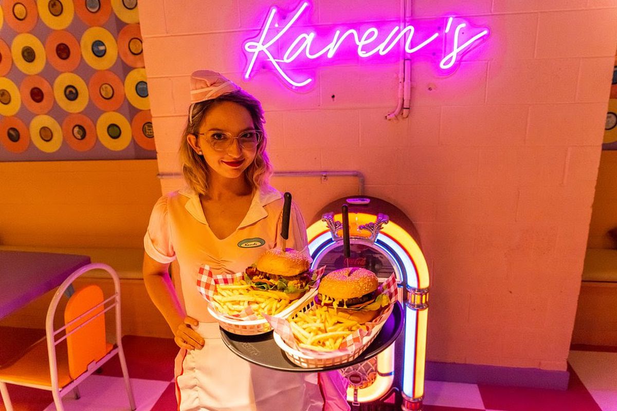 A person in a waitress outfit holding trays of burgers and fries in front of a neon jukebox.
