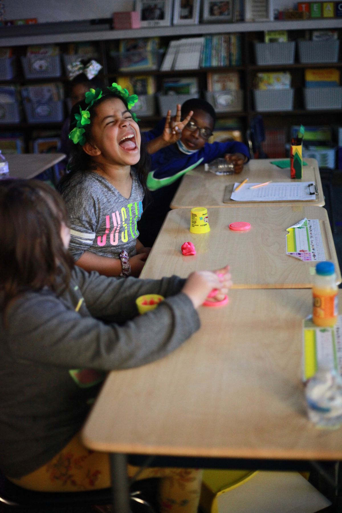 Three students are seated at their desks during an indoor recess.  A young girl in the middle shouts insistently as a boy next to her gestures with his fingers.