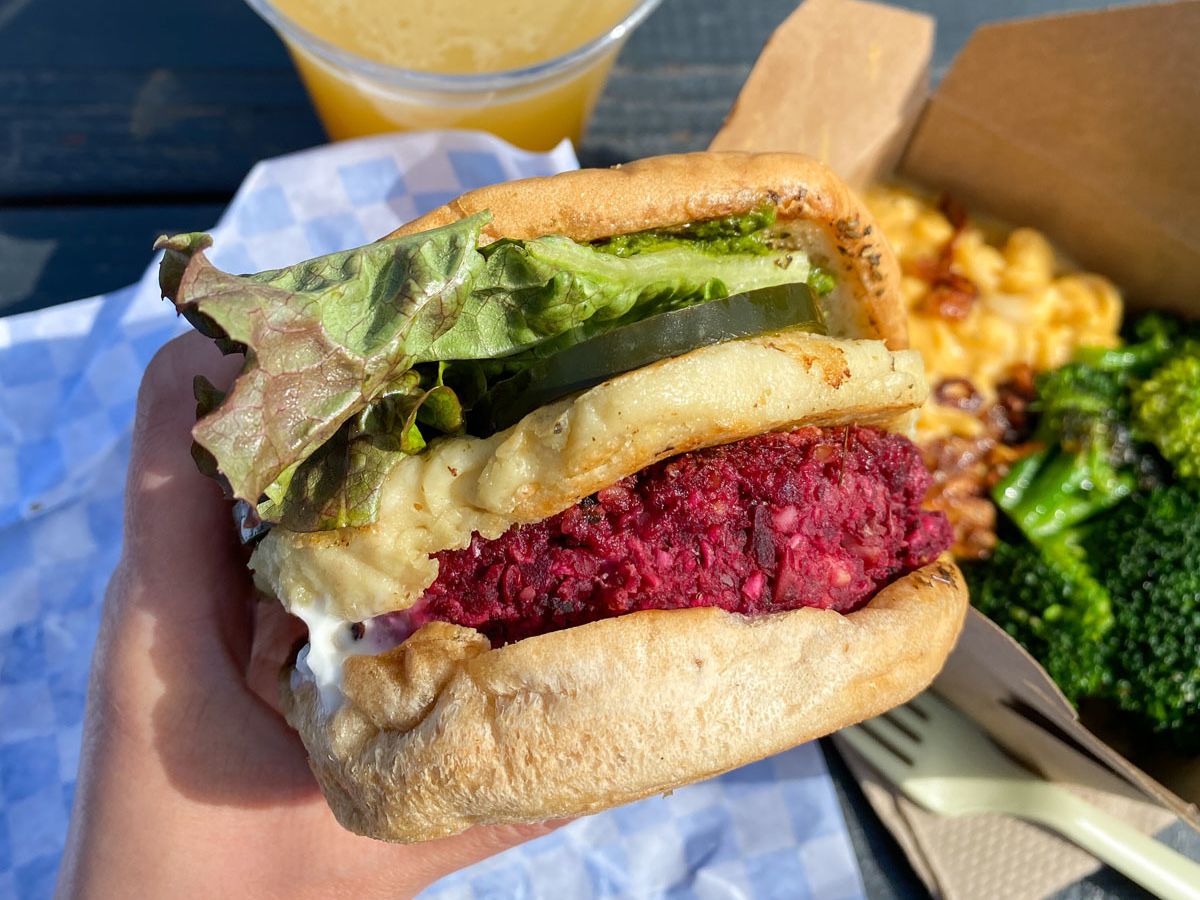 A photo of the beet, lentil, and walnut burger with cashew mozzarella from Flourish food truck, pictured next to a takeout container of mac and cheese and beer.