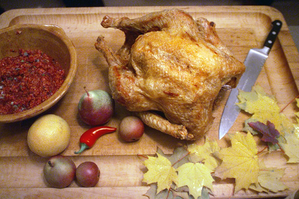 A turkey sits on a cutting board next to a knife, leaves, fruits and vegetables, and stuffing.