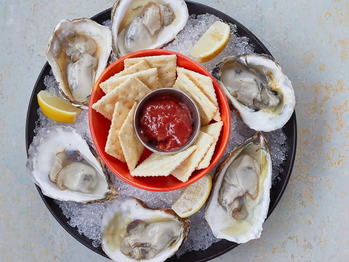 A plate filled with crushed ice topped with six oysters on the half shell with a small red bowl containing saltines and cocktails sauce in the center.