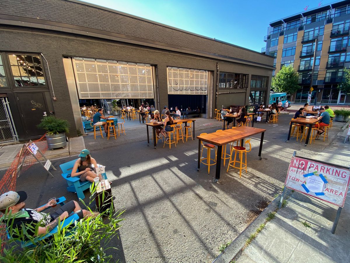 On a sunny day, several tables are set up outside Optimism Brewing on Broadway Court, with masked patrons sitting down
