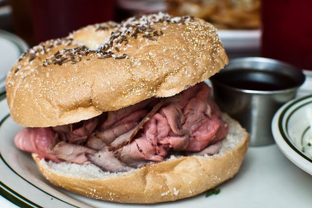 A beef on weck sandwich with rare roast beef on a kimmelweck bun and a silver cup of au jus