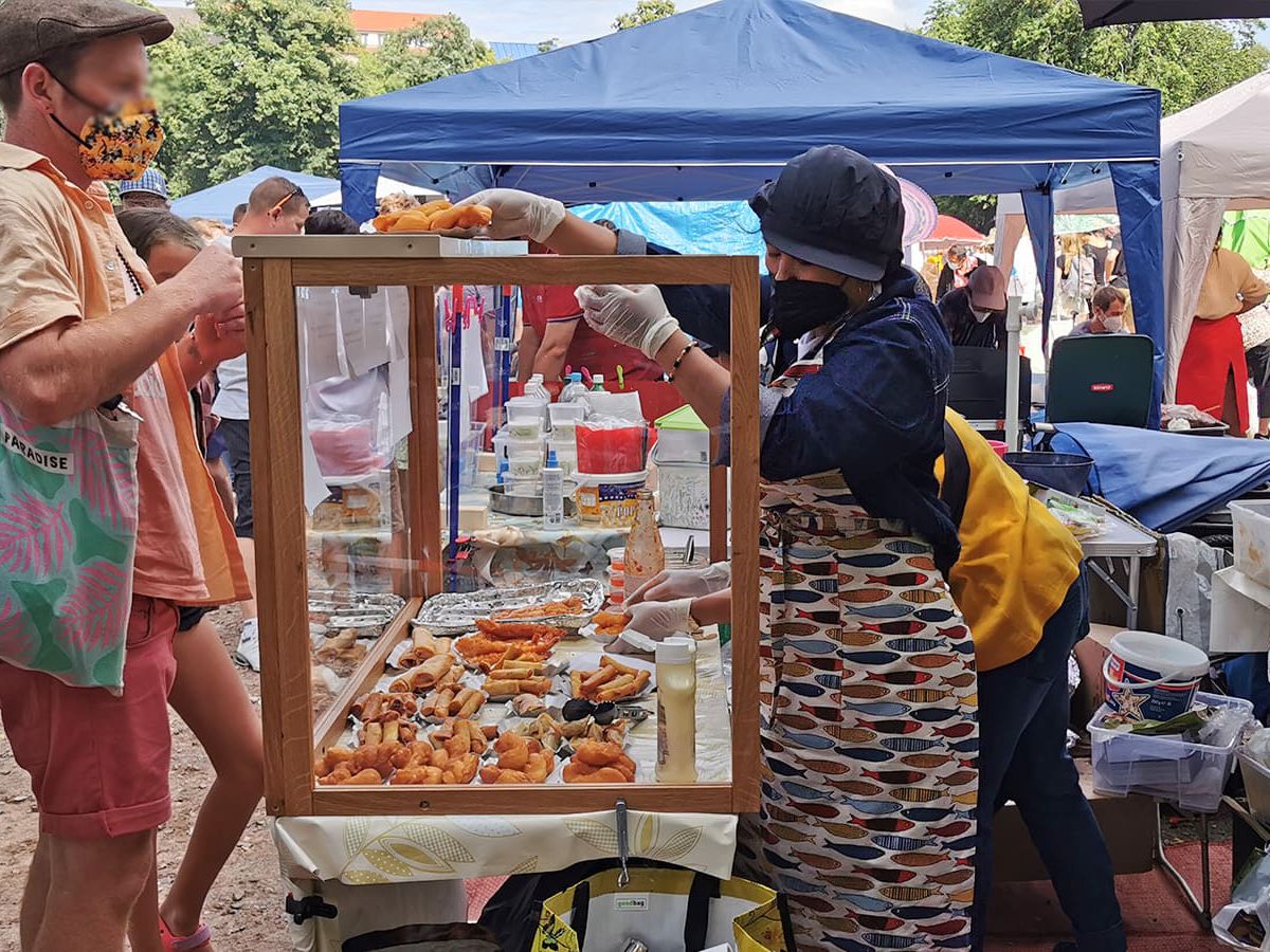 A vendor hands an item over the top of the glass case at a makeshift food stall in a sunny park to a customer waiting on the other side