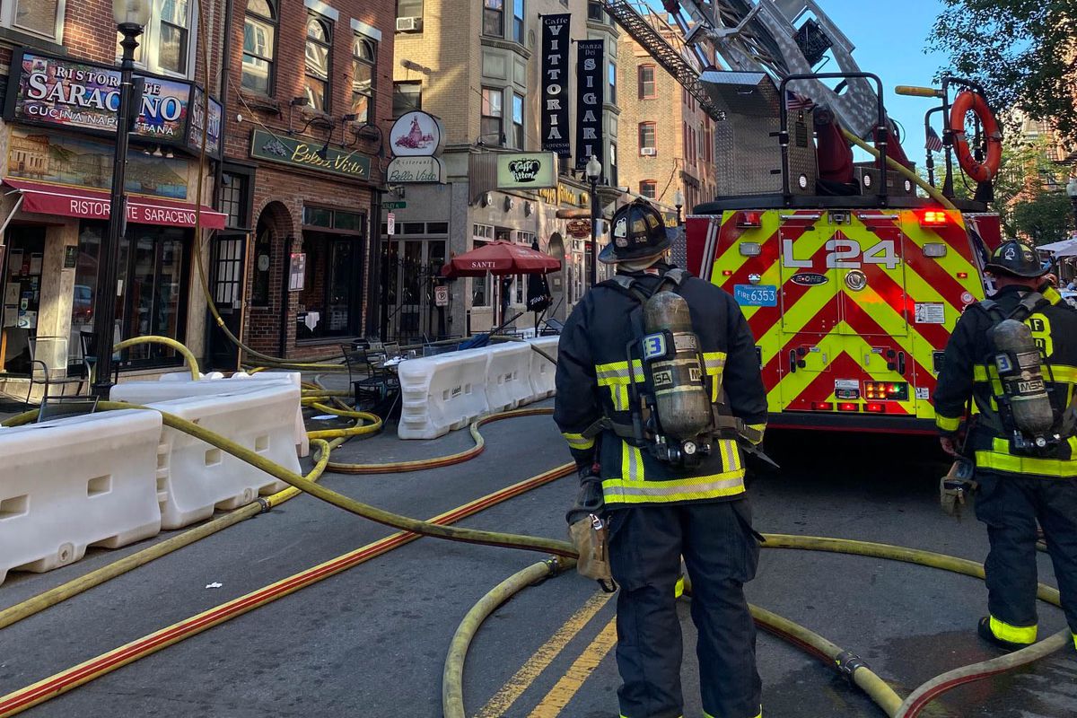 A fire truck and two firefighters are on a narrow city street with Italian restaurants lining the ground floors of several brick buildings.