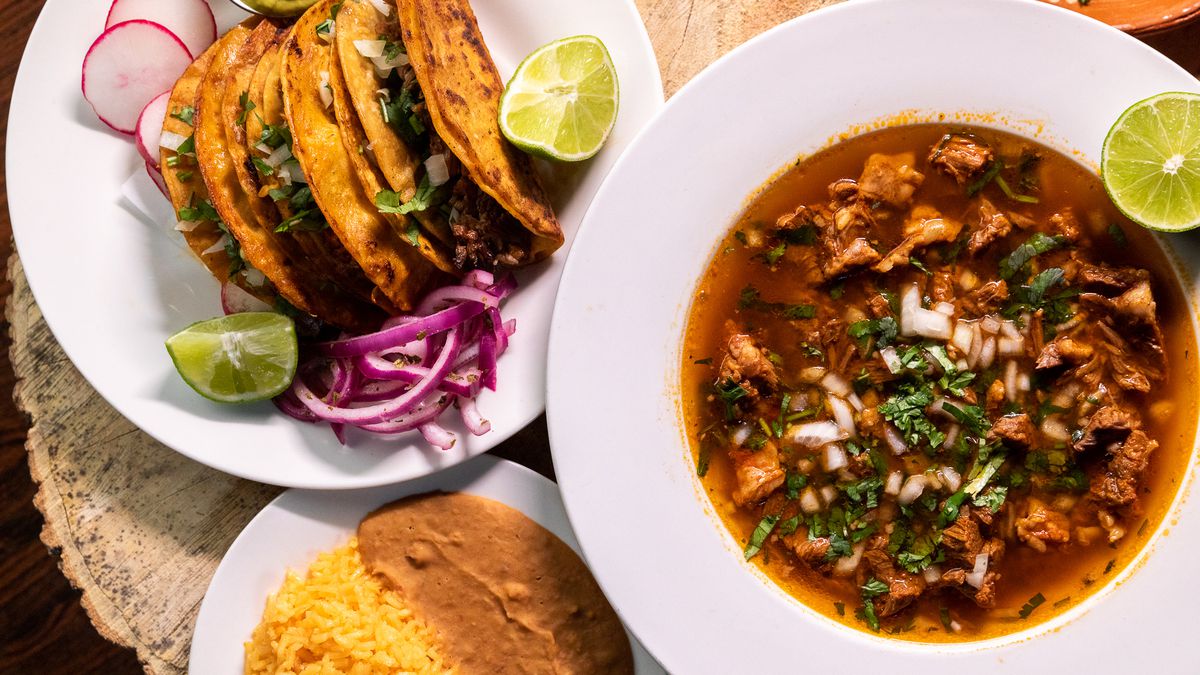 White plates of brick red birria stew and and crispy fried tacos surrounded by sliced limes, red onions, and slices of radish. 