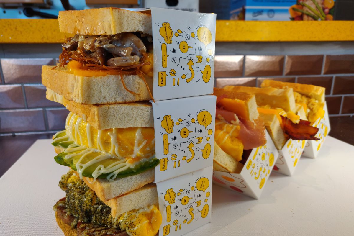 Three sandwiches on fluffy milk bread, oozing with cheese, overflowing with meat, stacked in cardboard sleeves beside paper boats of french fries