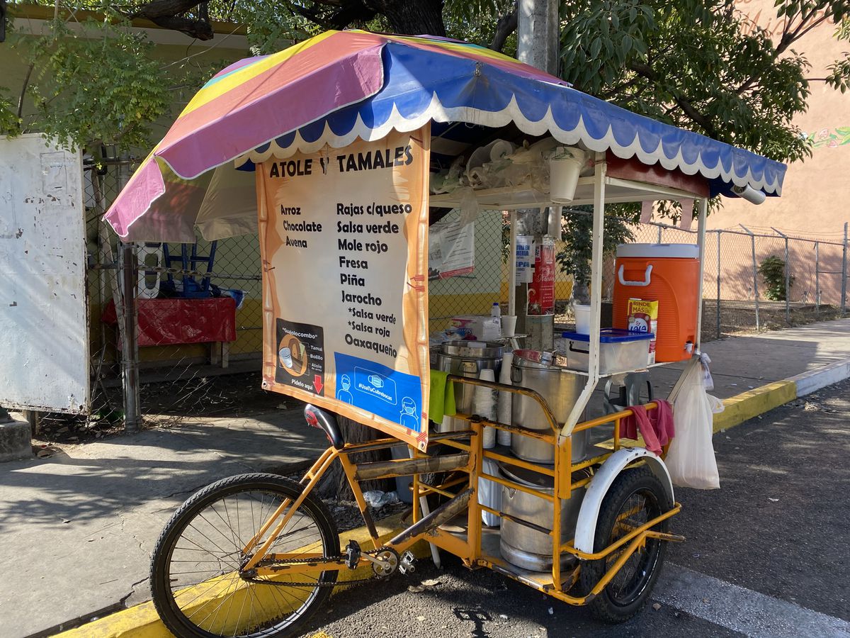 A tamal cart built over a bike, with a large colorful menu at the front, rainbow umbrella, cooler for serving drinks, and kitchen tools
