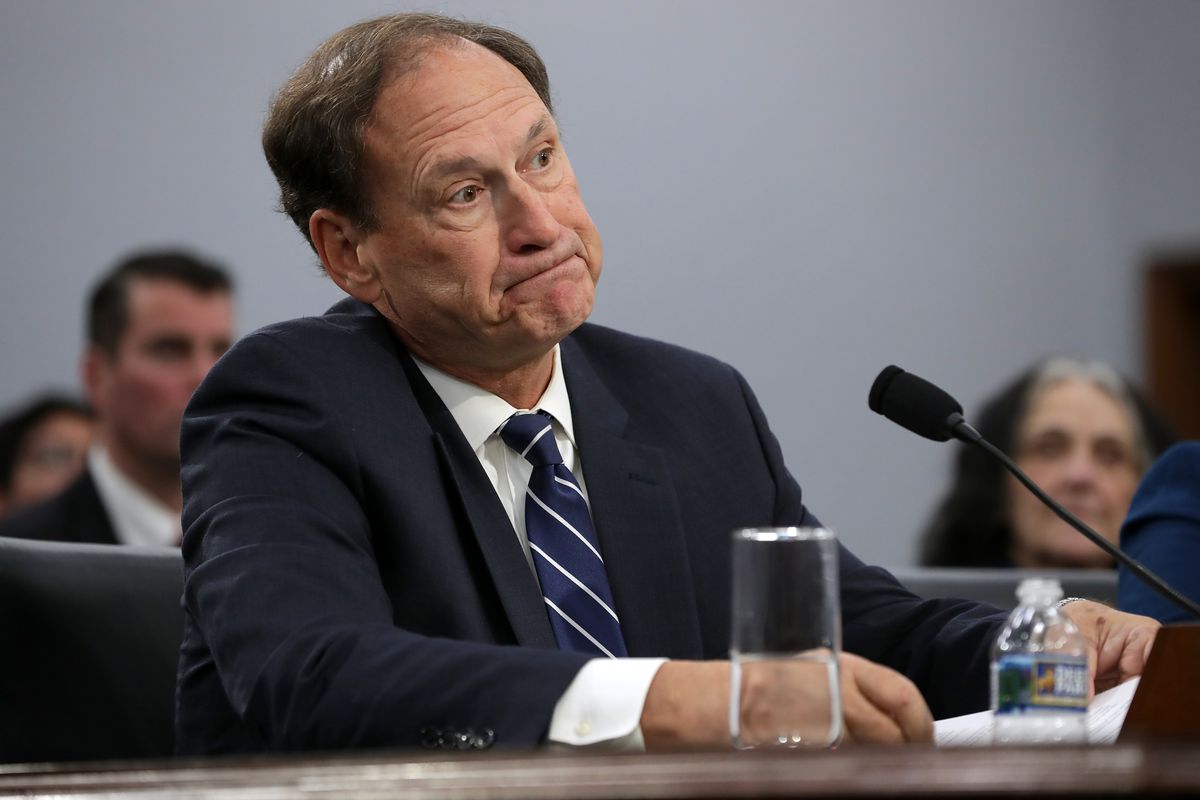 Samuel Alito sits at a desk facing a microphone. 