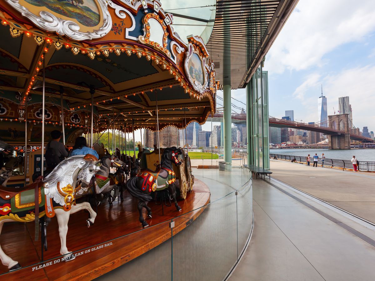 A carousel which has ornamental horses is next to a waterfront. In the distance is a bridge and a city skyline with buildings.