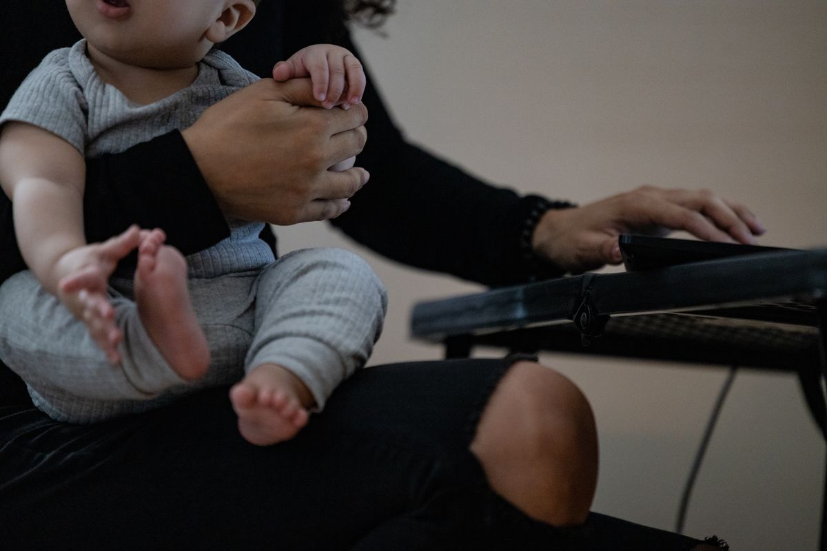 A woman holds her infant, who is wearing grey clothing, as the baby grasps her thumb and reaches for its foot as she works on a computer.