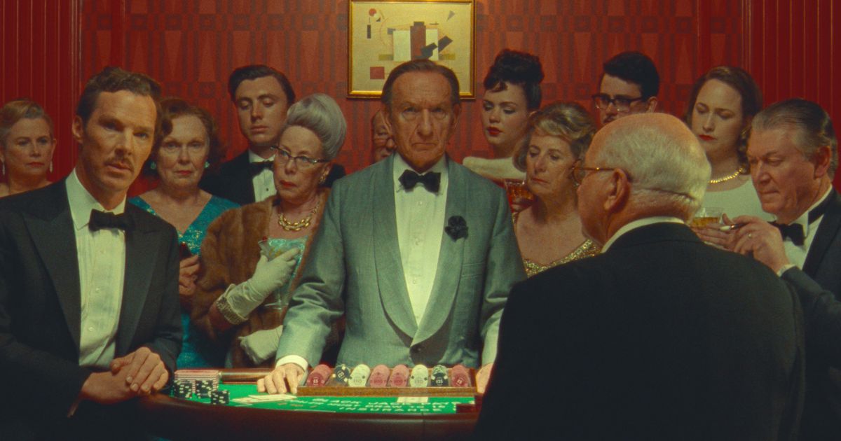 Benedict Cumberbatch (in a tuxedo as Henry Sugar) and Sir Ben Kingsley (as a croupier) look into the camera as they stand at a table in a casino, surrounded by a curious crowd of well-dressed people, in Wes Anderson’s Netflix film The Wonderful Story of Henry Sugar
