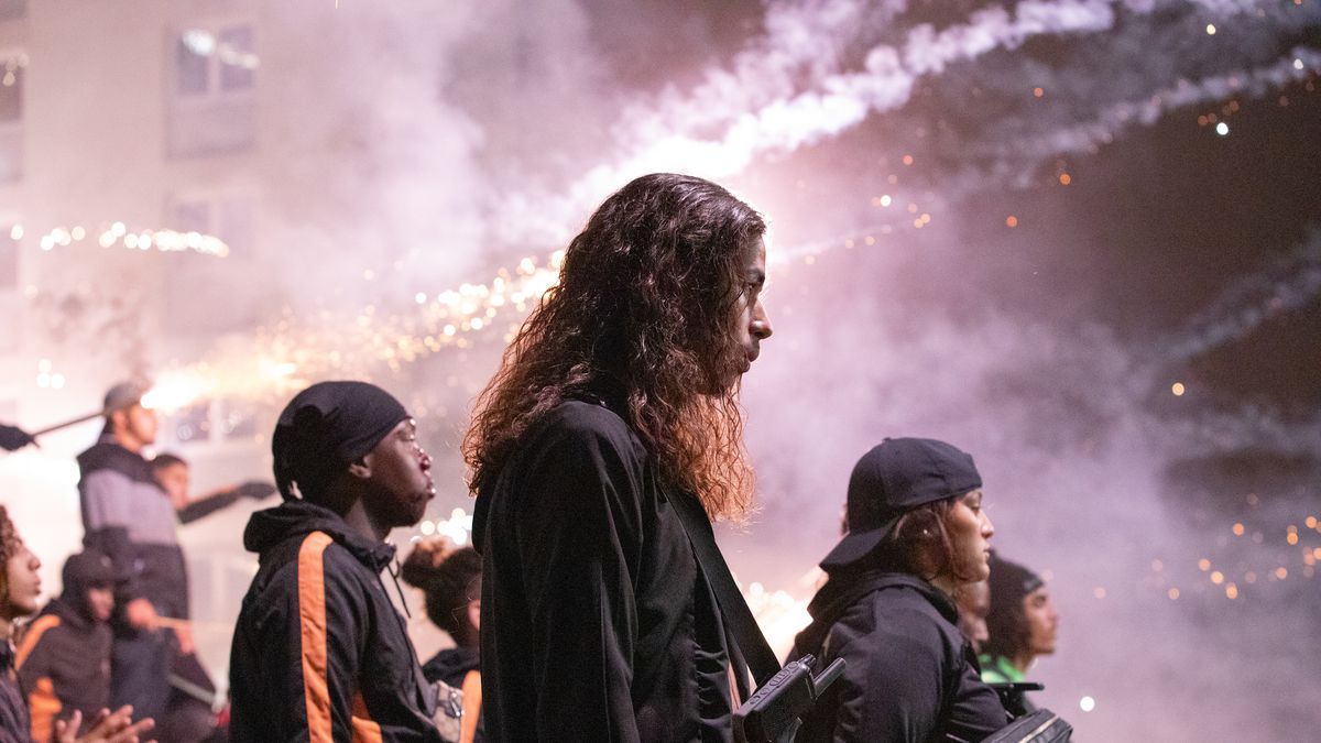 A side profile shot of a group of young men in black tracksuits with smoke and fireworks in the distance.