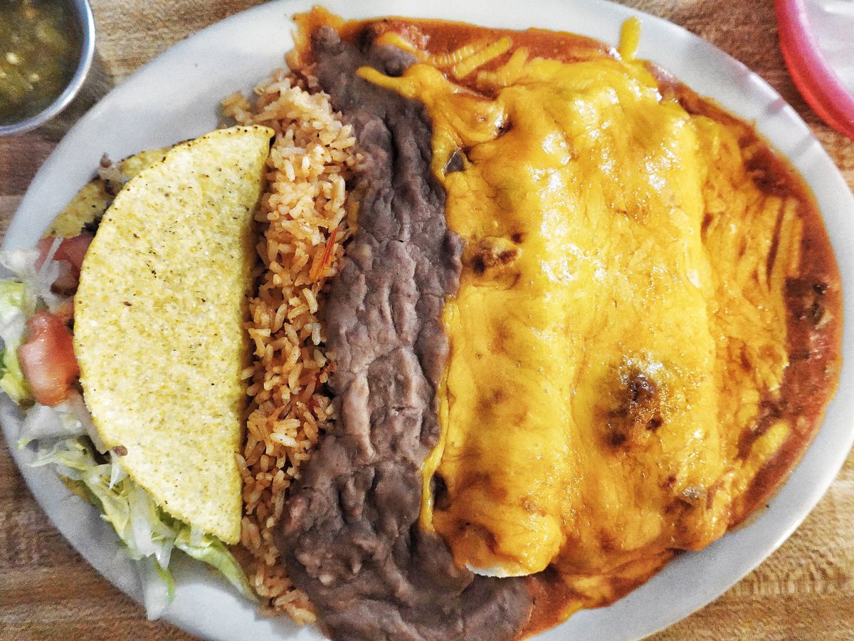 A white oval plate with a crispy taco, rice, refried beans, and enchiladas covered in yellow cheese sauce.