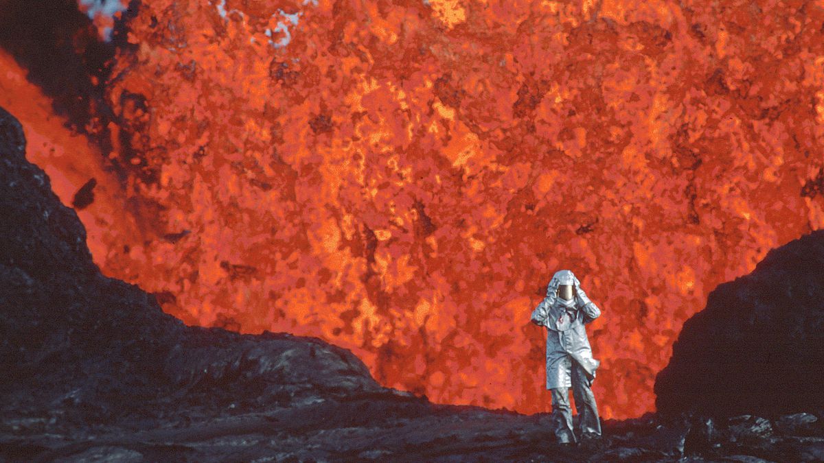 A person in a fire-proof suit walks away from the mouth of geyser overflowing with lava.