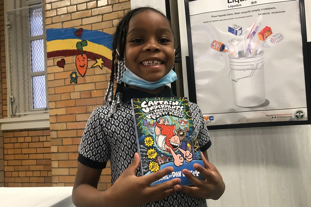 A little girl with braided hair smiles at the camera, posing with a copy of a Captain Underpants children’s book.
