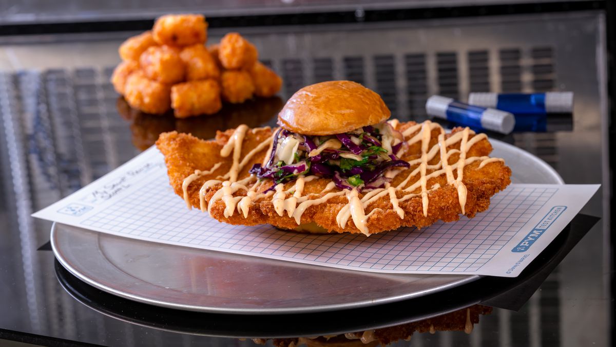 A giant fried chicken patty hangs out of the sides of a small bun sandwich with tater tots on the side