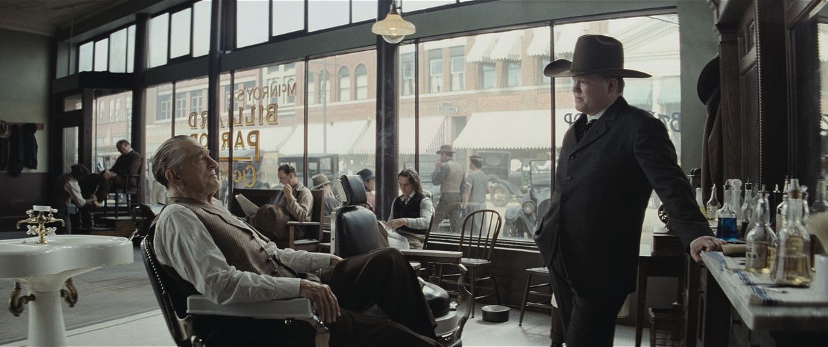 Jesse Plemons, in a huge cowboy hat, stands and interrogates Robert De Niro, sitting in a barber shop, in Killers of the Flower Moon