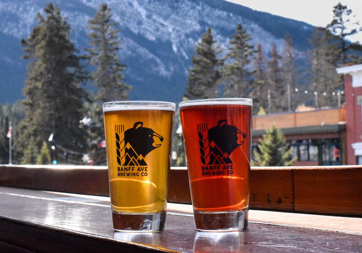 Two pints in branded glassware on an outdoor bar looking out on a city street, evergreen forest, and huge mountains