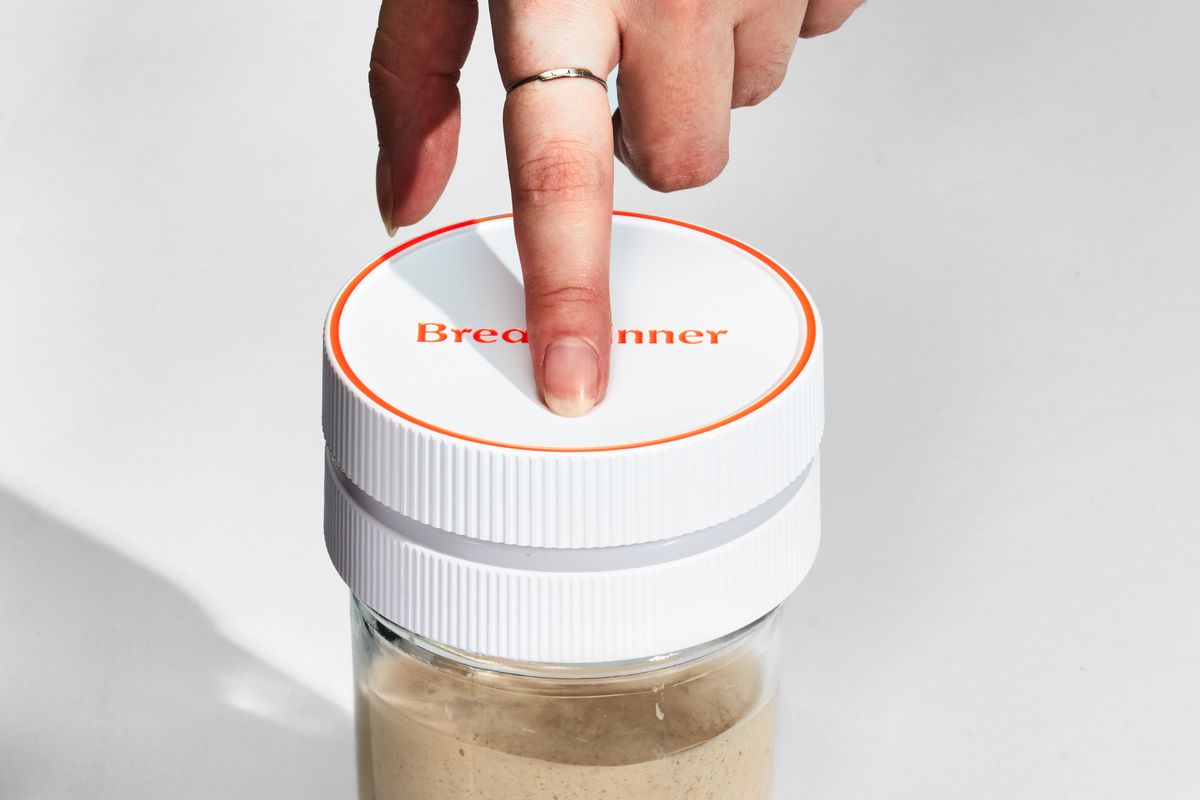 The Breadwinner device on top of a mason jar filled with starter. A person is clicking down on the lid to register a feeding.