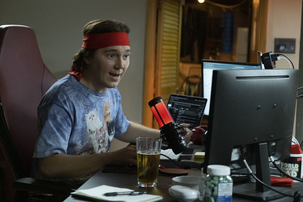 Paul Dano as Keith Gill, aka Roaring Kitty, sitting at a bank of monitors at his desk with a glowing mic in front of him, wearing a novelty T-shirt featuring fluffy kittens, and a red bandana tied around his head in Dumb Money