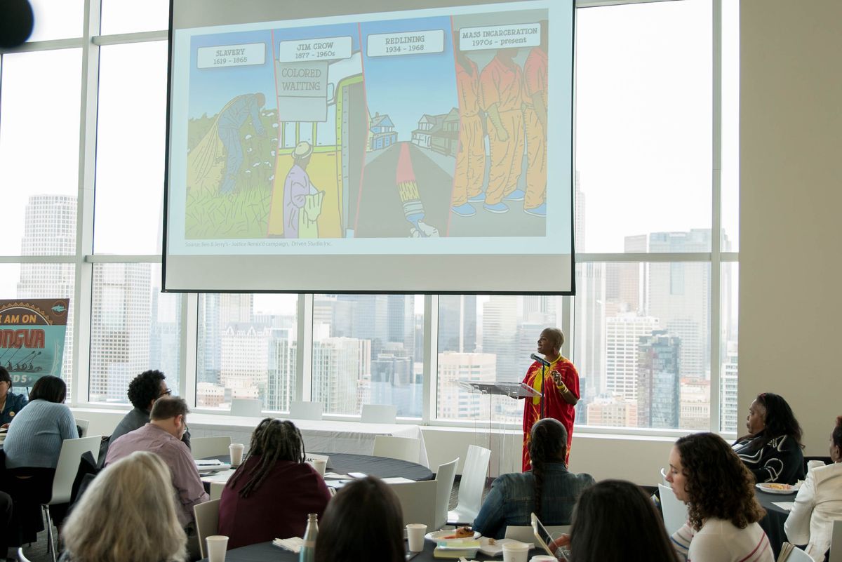 Against the backdrop of downtown LA, a woman stands in a conference room to give a presentation, with a slide that reads Slavery, Jim Crow, Redlining, and Mass Incarceration.