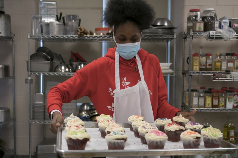 A young woman in a red Adidas sweatshirt carries a tray of cupcakes during a culinary arts class.
