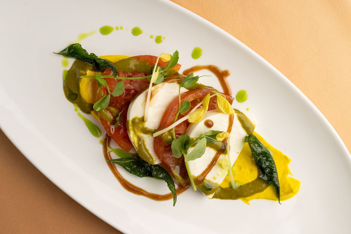 An oblong white plate topped with a caprese salad featuring half-moon slices of tomato and mozzarella with bright yellow turmeric oil and mint chutney.