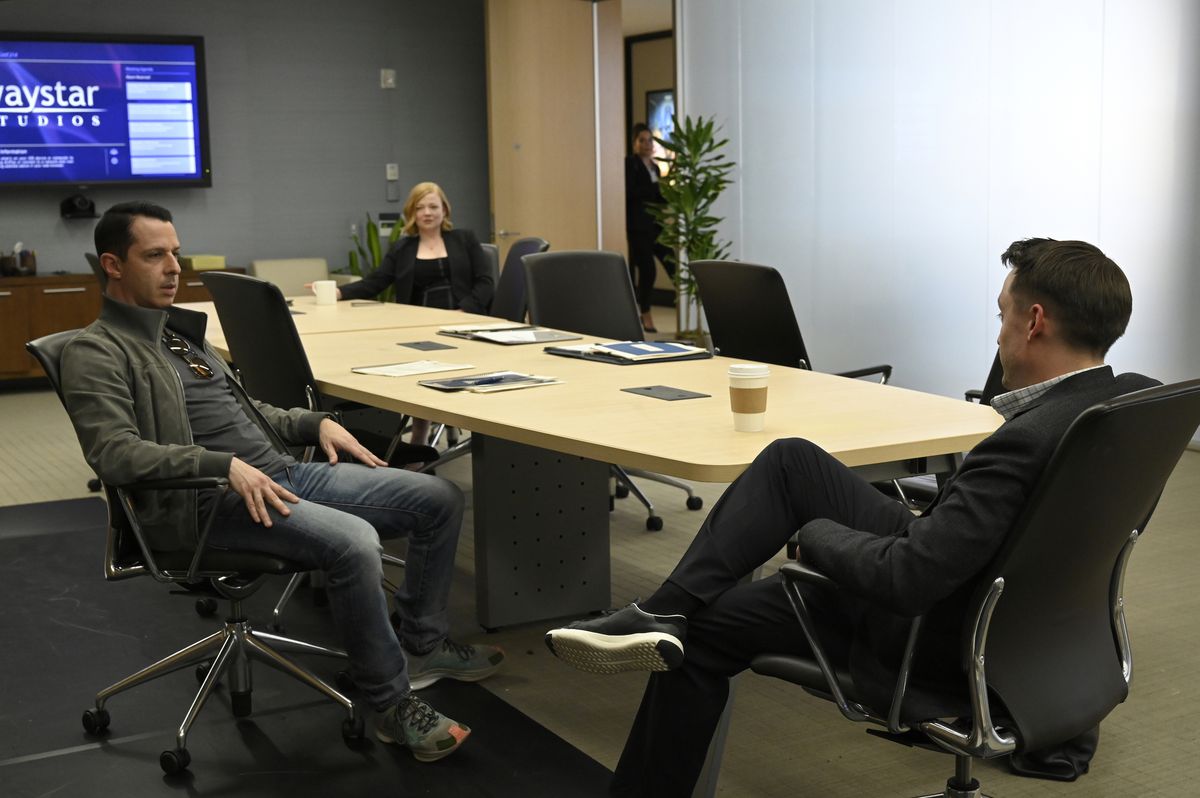 Shiv (Sarah Snook) sits on the far side in the background at a conference table while her brothers Kendall (Jeremy Strong) and Roman (Kieran Culkin) sit and talk closer to the foreground